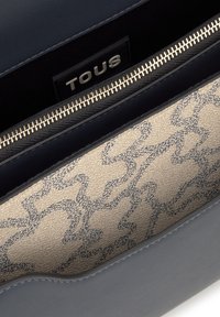 Primer plano del interior de un bolso de cuero azul que muestra un forro de tela beige con un patrón abstracto en negro y un zipper plateado con la etiqueta de la marca TOUS.