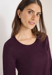 Burgundowa bluzka z długim rękawem i okrągłym dekoltem, wykonana z gładkiego materiału. Posiada dopasowany krój oraz minimalne detale szwów.