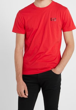T-shirt en coton rouge à manches courtes et col rond. Présente un petit logo EA7 en noir sur la poitrine. Porté avec un pantalon noir.