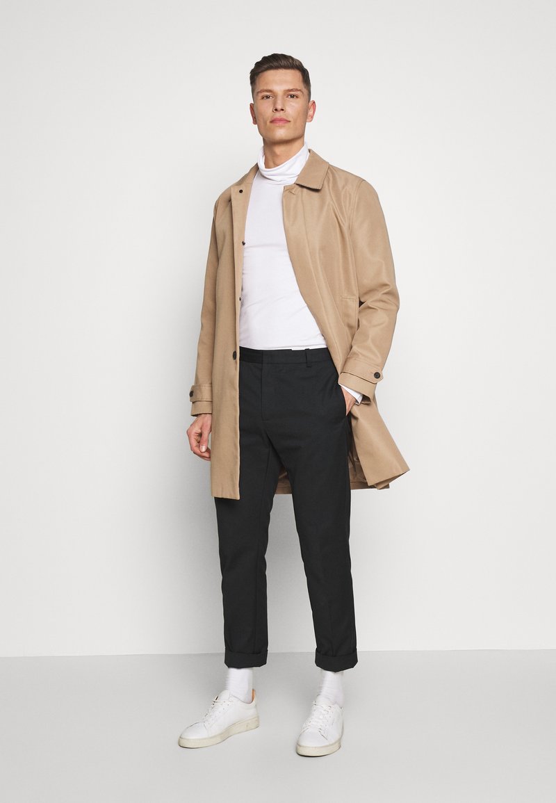 Gabardina ligera beige, camiseta blanca, pantalones negros ajustados y zapatillas blancas. Presenta un cierre de botones en la parte frontal y puños enrollados.