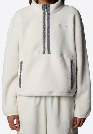 Person iført en hvid fleecepullover med halv lynlås og sidelommer, med The North Face-logo på brystet og bukser.