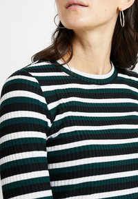 T-shirt à manches longues rayé avec des bandes horizontales noires, blanches et turquoise, doté d'une texture côtelée et d'un col rond.