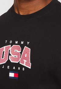 Black cotton T-shirt with round neckline, featuring bold red and white "TOMMY USA JEANS" text and a small multicolored brand logo at the bottom.