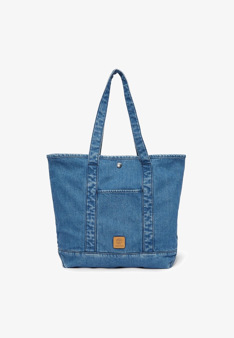 Sac fourre-tout en denim bleu avec longues anses, poche avant sécurisée par un bouton pression en métal, et petite étiquette de marque en cuir sous la poche.