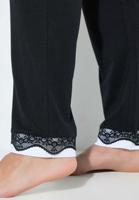 Zwarte broek van een gladde stof, met een witte zoom en zwart kanten rand aan de onderkant. Het ontwerp bevat subtiele textuurdetails.