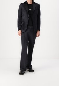 Mörk skräddarsydd kavaj med en knapp, tillsammans med mörka byxor. Kavajen har en jämn textur och slagkragar, bärs över en svart t-shirt.