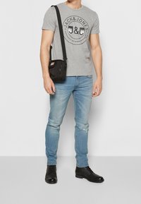 Camiseta de algodón gris con un logo negro, combinada con jeans desgastados de color azul claro y botas negras con cordones. Se lleva un pequeño bolso negro cruzado.