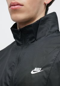 Fekete Nike dzsekit viselő személy, cipzárral zárt magas gallérral, valamint a fülön egy kis karika fülbevaló látható.