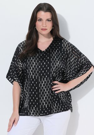 Donna con capelli lunghi e scuri che indossa una camicetta nera larga con motivi a tratti bianchi e pantaloni bianchi, che posa con una mano sul fianco.