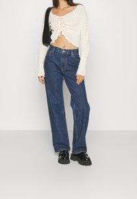 Pull en tricot court en crochet crème avec détails à nouer ; jeans en denim bleu foncé à jambes larges ; chaussures à plateforme épaisses noires.
