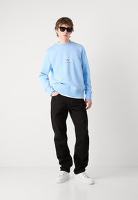 Felpa azzurra chiara con collo a giro e logo, abbinata a pantaloni neri e sneakers bianche, messa in risalto da occhiali da sole scuri.