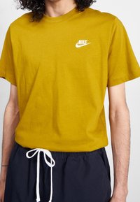 Hořčičkově žluté tričko Nike vyrobené z měkké bavlny s kulatým výstřihem. Obsahuje bílý potisk loga na hrudi. V kombinaci s námořnickými šortkami se šňůrkou.