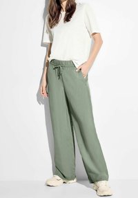Vrouw in lichtgroene wijde broek met trekkoord en witte sneakers, gecombineerd met een losse witte top met korte mouwen.