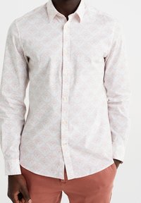 Camisa blanca con patrón floral en tonos rosas, de manga larga, con cuello abotonado y una tela texturizada. Presenta botones claros y un corte ajustado.
