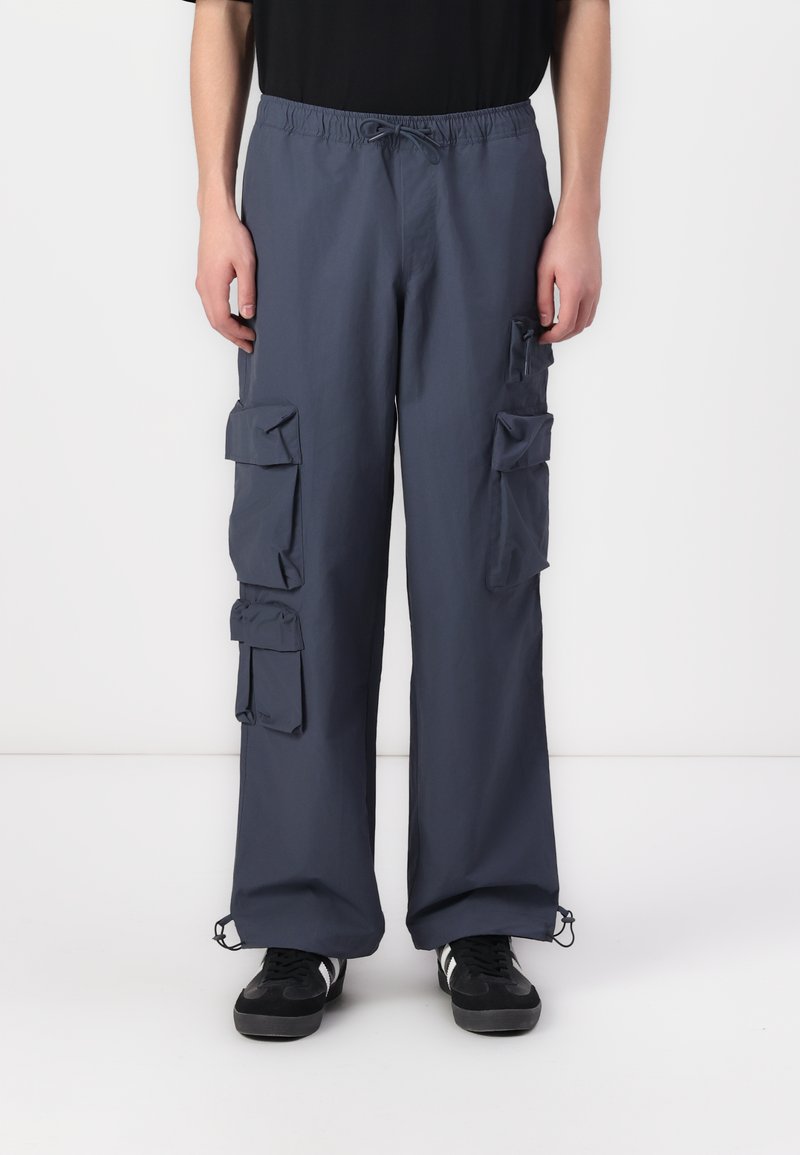 Navy cargo trousers with elastic waistband, multiple flap pockets, and drawstring hems. Made from lightweight fabric, featuring a relaxed fit.