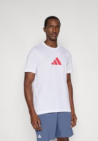 Biała bawełniana koszulka z czerwonym tr�ójkątnym nadrukiem i napisem na piersi, krótkimi rękawami i okrągłym dekoltem, zestawiona z niebieskimi szortami.