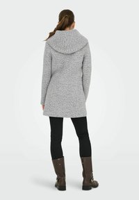 Femme avec une queue de cheval brune portant un manteau à capuche gris, des leggings noirs et des bottes brunes mi-mollet, se tenant de dos sur un fond blanc.