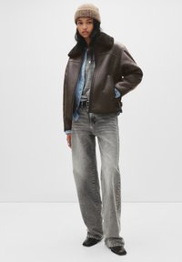 Jonge vrouw met een beige gebreide muts, donkerbruine shearling jas, gestreept blauw overhemd, grijze trui, wijde versleten jeans en zwarte puntige schoenen.