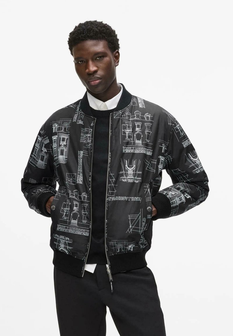 Jeune homme portant une veste bomber noire avec des croquis architecturaux blancs, un pantalon noir et une chemise blanche sous un pull noir.