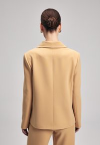 Beige přizpůsobený blejzr s klopovými límci, strukturovanými rameny a hladkým materiálem. Má čistý design zadní části, přičemž jsou vidět odpovídající kalhoty.