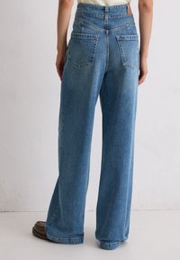 Persoon gekleed in loszittende blauwe denim jeans en bruine schoenen, staand tegen een effen lichtgekleurde muur als achtergrond.