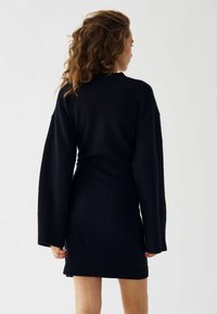 Melnā adīta kleita ar garām, plašām piedurknēm un ribotām malām. Piedāvā brīvu piegriezumu un vidukļa garumu, izceļoties ar maigu tekstūru.