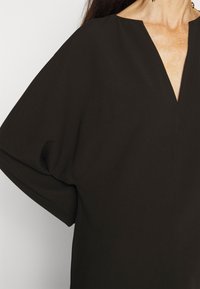 Kvinde iført en løstsiddende sort V-hals top med vide ærmer og øredobber i guldkæde, der viser hals og delvist bryst.