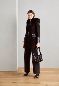 Cappotto nero con cappuccio in pelliccia sintetica e dettagli in pelle, chiusura con zip, due tasche con zip. Abbinato a pantaloni larghi scuri e una borsa nera.