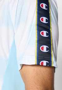 Sportshirt met korte mouwen in lichtblauw en wit, met een stippenpatroon en een zwarte zijaccent met rode en blauwe logo-accenten.