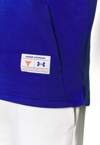 Maglietta sportiva blu con etichetta che presenta il logo Under Armour e un design di un toro arancione. Il tessuto è strutturato e presenta uno spacco laterale per facilitare il movimento.