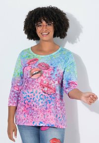 Lächelnde Frau mit lockigem Haar, die eine klare rosa Brille, ein farbenfrohes Oberteil mit Lippen-Prints und Jeans mit roten Lippenaufnähern trägt.