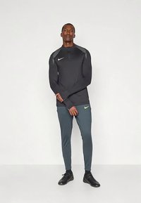 Melnā garroku sporta virsdrēbe ar pelēkiem akcentiem, rāvējslēdzēja kakla izgriezums, kopā ar pieguļošām tumši pelēkām biksēm un melniem apaviem. Redzamas Nike logos.