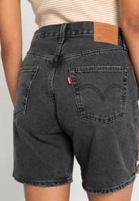 Shorts de mezclilla negra con una cintura alta, dobladillo deshilachado y dos bolsillos traseros. Presenta una etiqueta de cuero marrón con el logo de la marca en la cinturilla.