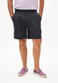 Shorts plissés bleu marine foncé en tissu léger, dotés de poches latérales et d'une taille épurée. Portés avec des chaussures à enfiler violettes.