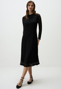 Schwarzes Spitzenkleid mit langen, durchsichtigen Spitzenärmeln, rundem Halsausschnitt und ausgestelltem Schnitt. Abgerundet mit einem gewellten Saum.