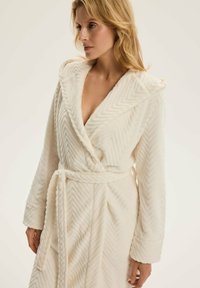 Biała szata z wzorem w zygzak, wykonana z pluszowego materiału, z kapturem, długimi rękawami i związanym paskiem w talii.