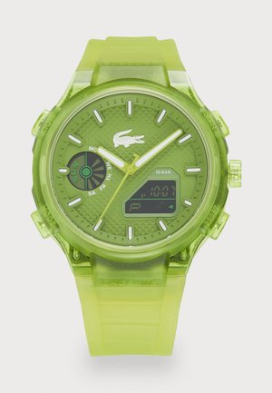 Montre numérique verte avec un bracelet en caoutchouc transparent, un cadran texturé, des accents blancs et un affichage numérique. Présente le logo Lacoste et plusieurs cadrans.