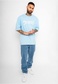 Lyseblå oversized t-shirt med hvid grafisk tekst, parret med løse blå denim jeans og hvide sneakers. Enkel, afslappet design.