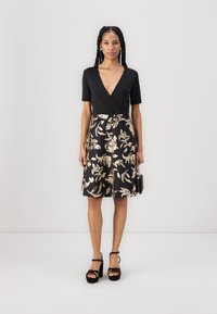 Top noir à manches courtes, associé à une jupe mi-longue à motifs avec des feuilles dorées sur un fond noir. Sandales à talons noires.