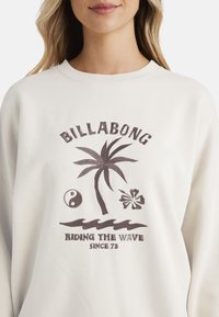 Sudadera color crema con gráficos impresos en marrón: una palmera, símbolo del yin-yang, flor y texto "BILLABONG MONTANDO LA OLA DESDE EL 73."