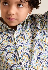 Een lichtblauwe overhemd met een bladerenpatroon in blauw, groen en geel op een witte achtergrond. Voorzien van een kraag en gele knopen.