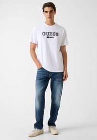 Biała koszulka z krótkim rękawem z czarnym logo "GUESS" i haftem "EST. 1981". Noszona z wyblakłymi, niebieskimi, prostymi jeansami i jasnymi sneakersami.