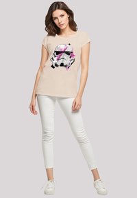 Hellbeiges T-Shirt mit einem schwarzen und pinkfarbenen Aufdruck eines Sturmtruppler-Helms, kombiniert mit weißen Skinny-Jeans und weißen Sneakern.