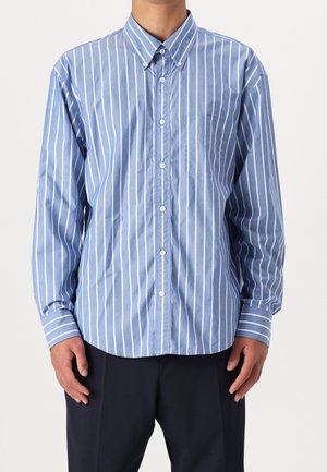 Mies, joka käyttää sinistä pitkähihasta nappipaitaa, jossa on valkoiset pystysuorat raidat ja rintatasku, yhdistettynä tummiin housuihin, yksinkertaisen taustan edessä.