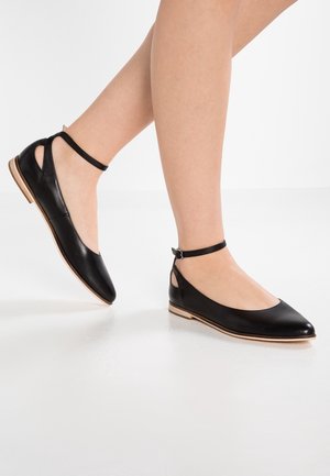 Jambes portant des chaussures plates pointues en cuir noir avec des brides autour de la cheville, posées sur un sol blanc réfléchissant.