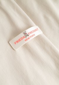 Witte stoffen label op beige stof met rode tekst "Frieda & Freddies New York" en een kleine zilveren hondenbedel eraan bevestigd.