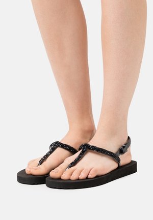 Sandales en caoutchouc noires avec un dessus texturé orné de perles, semelle plate en caoutchouc et bride ajustable à la cheville pour un bon maintien.