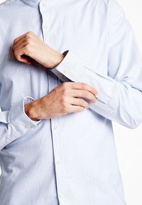 Camicia a righe blu chiaro con colletto e polsini abbottonati. Il tessuto appare liscio, con una consistenza croccante e righe verticali ben evidenti.