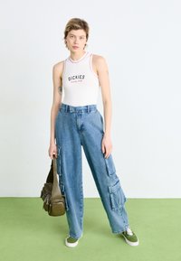 Balts bodijs ar "DICKIES" logo, kombinēts ar gaiši ziliem plašiem kravas džinsiem, kuriem ir vairāki kabatas. Modelis tur zaļu rokassomu.