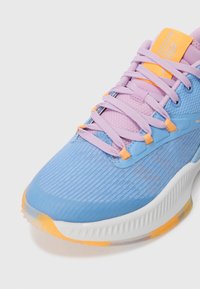 Gros plan sur une chaussure de sport bleu et lavande avec des accents orange, des lacets violets et une semelle blanche avec des détails orange.
